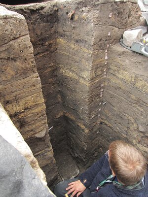 Foto från utgrävningen där man ser olika lager i marken. En person sitter och tittar på dem.