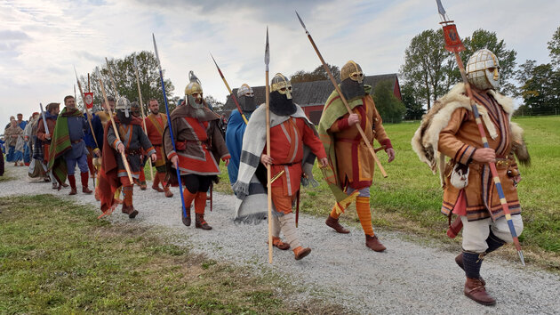 Människor utklädda till forntida rigare med röda och bruna dräkter, hjälmar och spjut